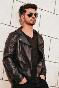 Young man stands against wall wearing leather jacket and sunglasses